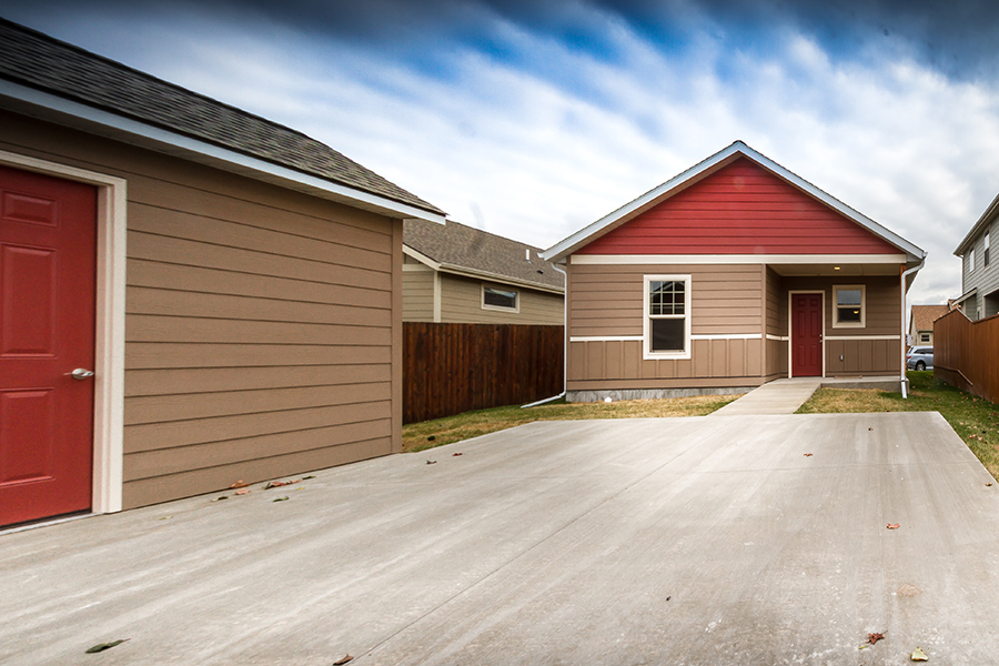 A sidewalk connects a concrete driveway pad to the porch of a tan and red Visitable home. Off the driveway sits a matching shed with a zero-step threshold and silver lever-style door handle.